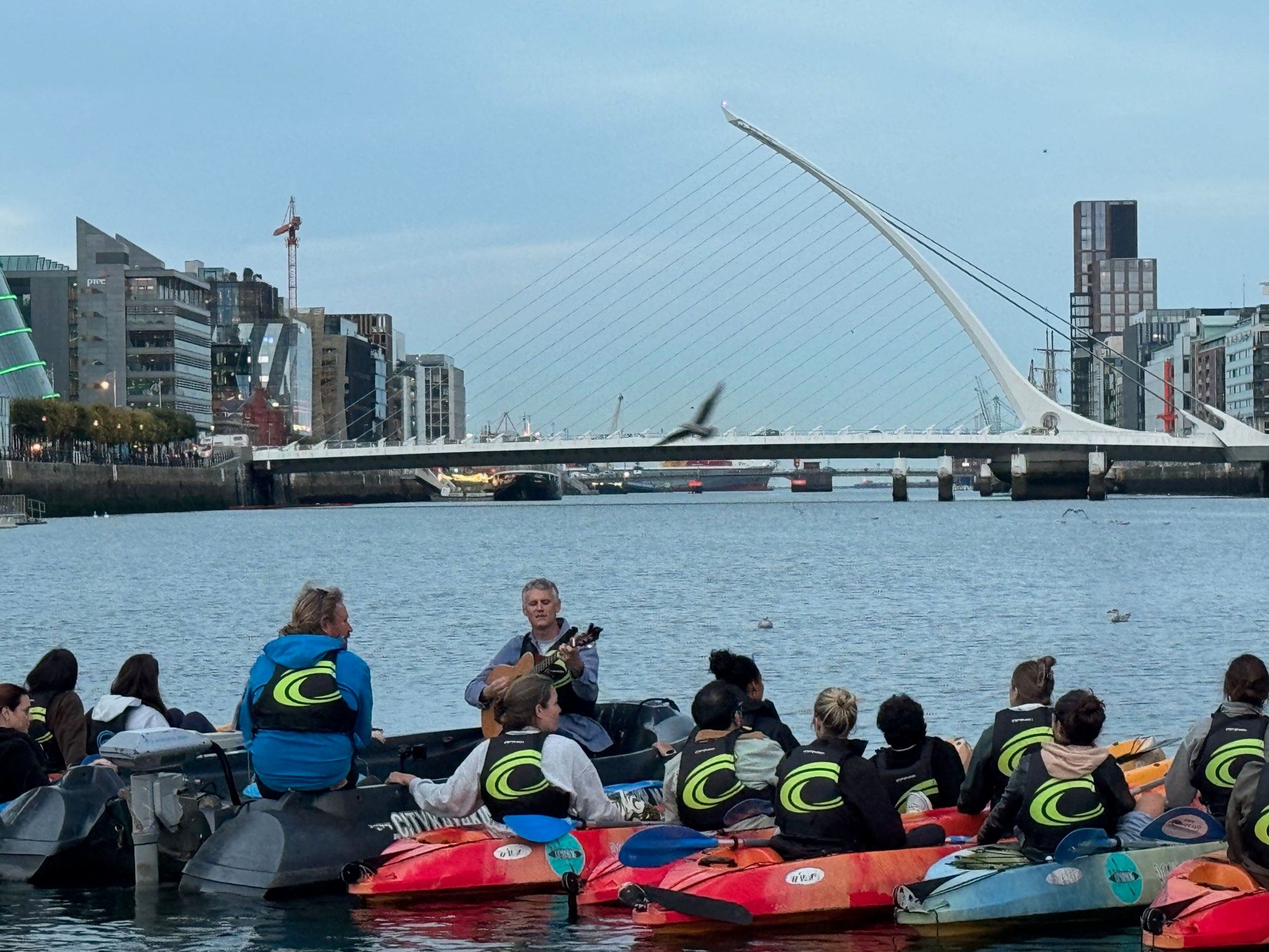 A musician playing guitar from a support boat as the kayak group rafts up under Samuel Beckett Bridge